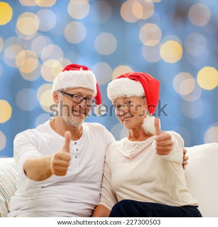 family, holidays, christmas, age and people concept - happy senior couple in santa helper hats sitting on sofa and showing thumbs up gesture over blue lights background