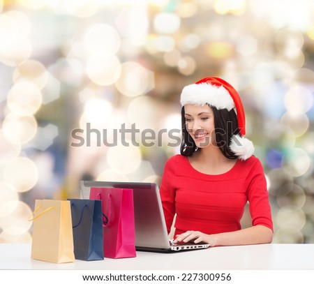 christmas, holidays, technology and people concept - smiling woman in santa helper hat with shopping bags and laptop computer over lights background