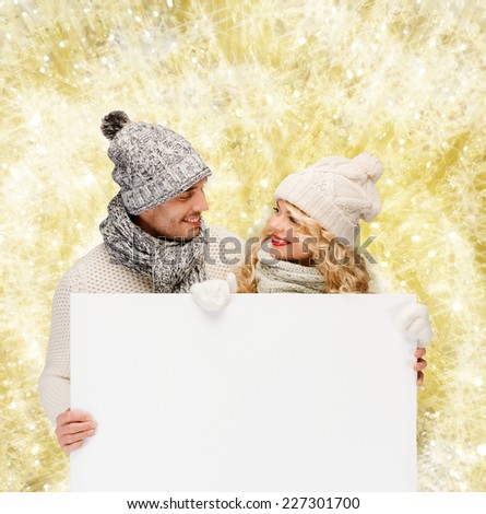 winter, holidays, christmas, advertisement and people concept - smiling couple in winter clothes with white blank billboard over yellow lights background