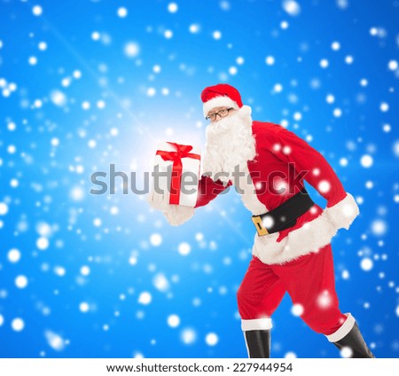 christmas, holidays and people concept - man in costume of santa claus running with gift box over blue snowy background