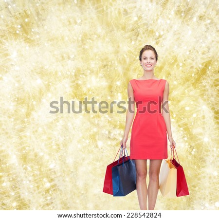 shopping, sale, christmas and holidays concept - smiling elegant woman in red dress with shopping bags over yellow lights background