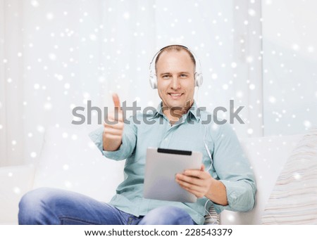 technology, people, winter, gesture and leisure concept - smiling man in headphones with tablet pc sitting on couch and showing thumbs up at home