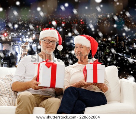 family, holidays, christmas, age and people concept - happy senior couple in santa helper hats with gift boxes over snowy night city background