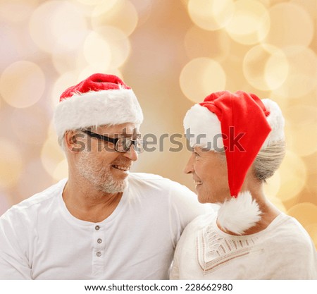 family, holidays, christmas, age and people concept - happy senior couple in santa helper hats sitting on sofa over beige lights background