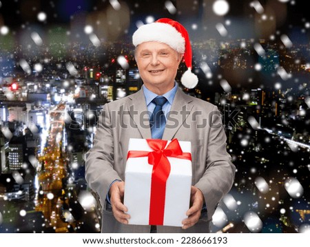 business, christmas, presents and people concept - smiling senior man in suit and santa helper hat with gift over snowy night city background