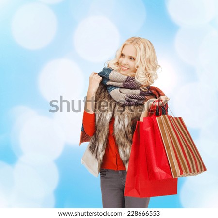 happiness, winter holidays, christmas and people concept - smiling young woman in winter clothes with red shopping bags over blue lights background