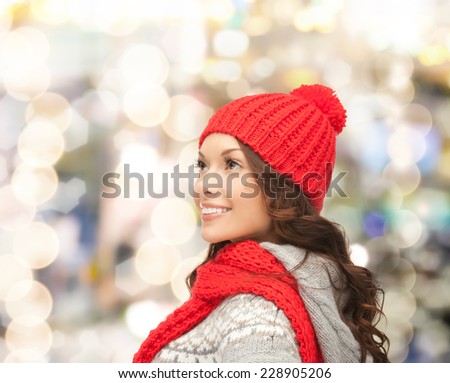 happiness, winter holidays, christmas and people concept - smiling young woman in red hat and scarf over lights background