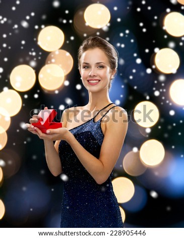 holidays, christmas, presents, luxury and people concept - smiling woman in dress holding red gift box night lights and snow background