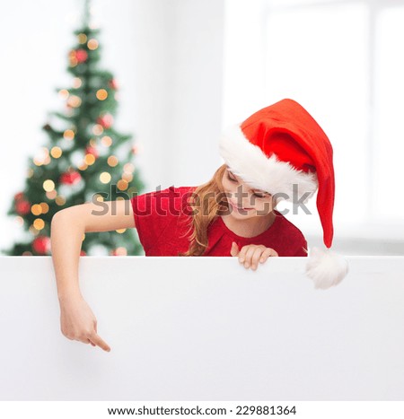 christmas, holidays and advertisement concept - happy girl in santa helper hat pointing finger on blank white board over living room with christmas tree background