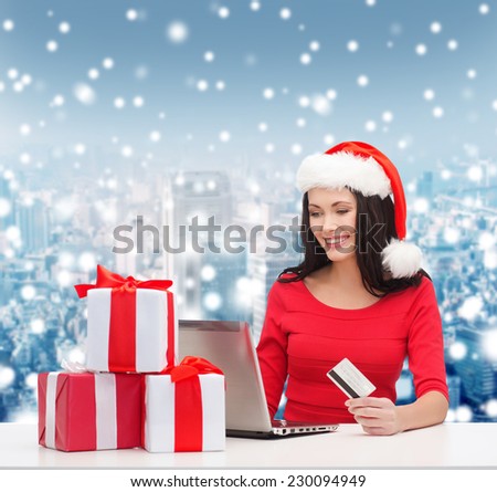 christmas, shopping and people concept - smiling woman in santa helper hat with gift boxes, laptop computer and credit card over snowy city background