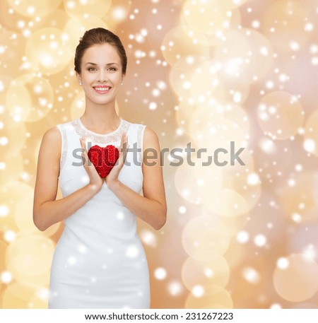 happiness, health, charity and love concept - smiling woman in white dress with red heart over beige background over beige lights background and snow