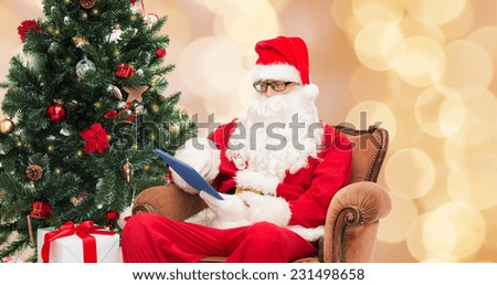 technology, holidays and people concept - man in costume of santa claus with tablet pc computer, gifts and christmas tree sitting in armchair over beige lights background