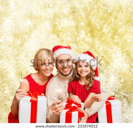christmas, holidays, family and people concept - happy mother, father and little girl in santa helper hats with gift boxes and sparklers over yellow lights background