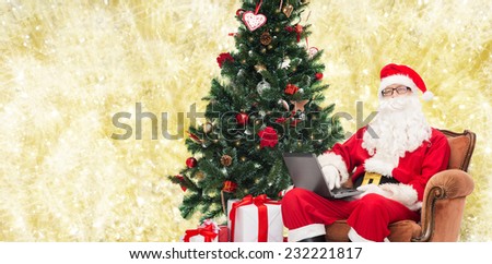 technology, holidays and people concept - man in costume of santa claus with laptop computer, gifts and christmas tree sitting in armchair over yellow lights background