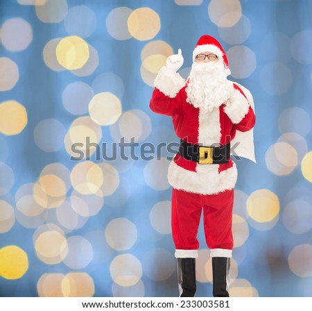 christmas, holidays, gesture and people concept - man in costume of santa claus with bag pointing finger up over blue lights background