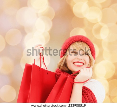 happiness, winter holidays, christmas and people concept - smiling young woman in hat and scarf with red shopping bags over beige lights background