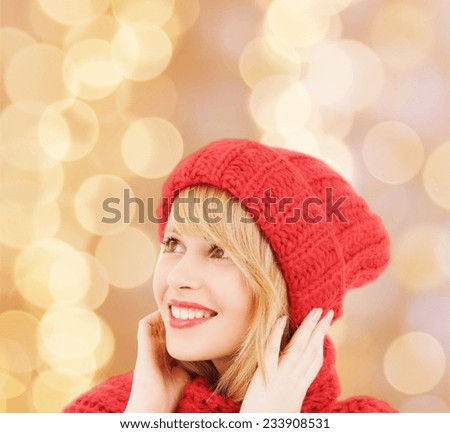happiness, winter holidays, christmas and people concept - smiling young woman in red hat and scarf over beige lights background
