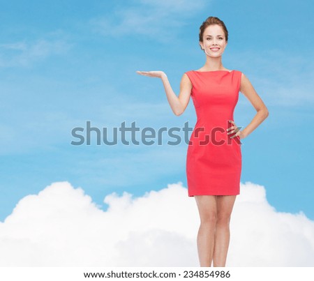 advertising and people concept - smiling young woman in red dress holding something on palm of her hands over blue sky and cloud background