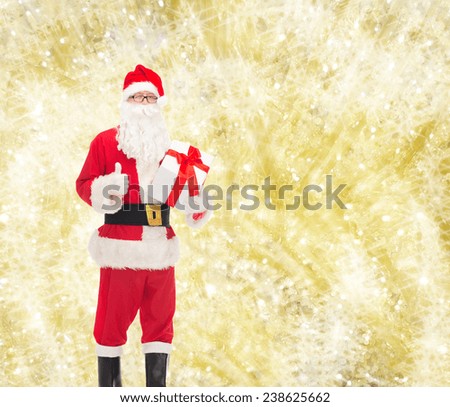 christmas, holidays and people concept - man in costume of santa claus with gift box showing thumbs up gesture over yellow lights background