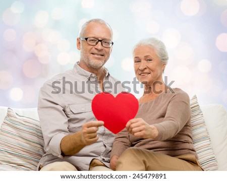 family, holidays, age and people concept - happy senior couple holding little red paper heart shape cutout and sitting on sofa over blue lights background