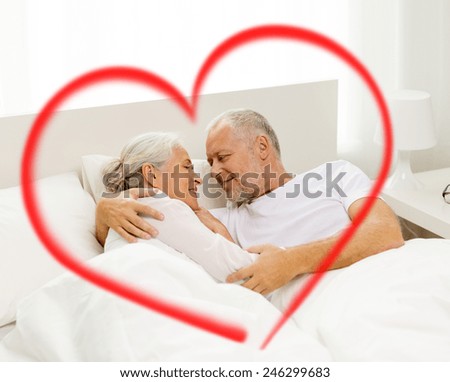 family, bedtime, valentines day, love and people concept - happy senior coupler lying in bad and hugging at home with big red heart shape