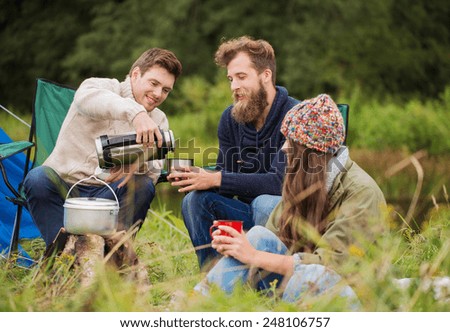 adventure, travel, tourism and people concept - group of smiling friends cooking food in dixie sitting around bonfire outdoors