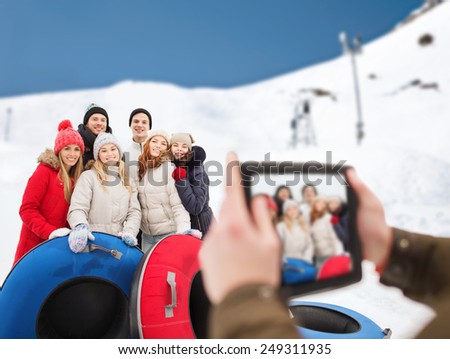 winter, leisure, sport, friendship and people concept - group of smiling friends with snow tubes taking picture by tablet pc computer over mountain background