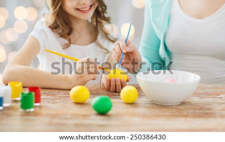 easter, family, holiday, people and childhood concept - close up of happy girl and mother with brushes coloring easter eggs over lights background