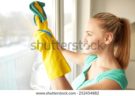 people, housework and housekeeping concept - happy woman in gloves cleaning window with rag and cleanser spray at home