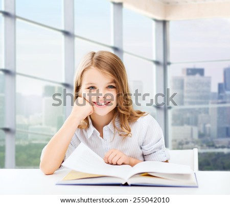 education, people, children and school concept - little student girl sitting at table with books and writing in notebook over classroom background