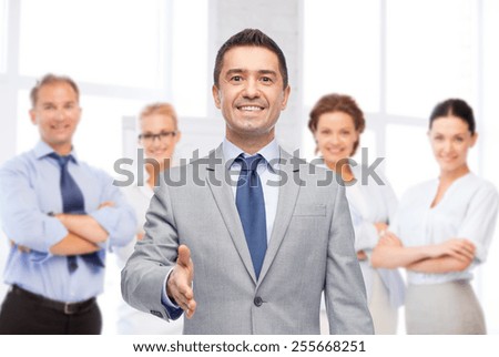 business, people, gesture, partnership and greeting concept - happy smiling businessman in suit with team over office room background shaking hand