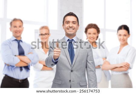business, people, gesture and office concept - happy businessman with team over office room background showing thumbs up