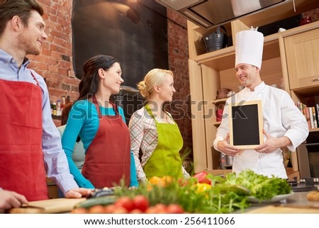 cooking class, culinary, food and people concept - happy friends and chef cook with blank menu chalk board in kitchen