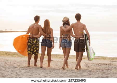 friendship, sea, summer vacation, water sport and people concept - group of smiling friends wearing swimwear with surfboards on beach from back