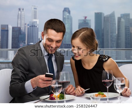 technology, food, holidays and people concept - smiling couple with smartphone eating at restaurant over city background