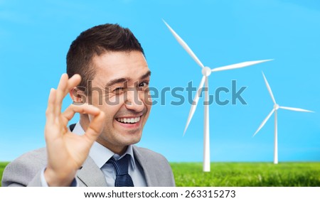 business, people, alternative energy and development concept - happy smiling businessman in suit showing ok hand sign over blue sky and windmills background