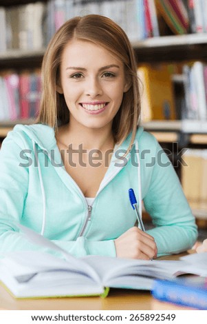 people, knowledge, education and school concept - happy student gir with book and pen writing in library