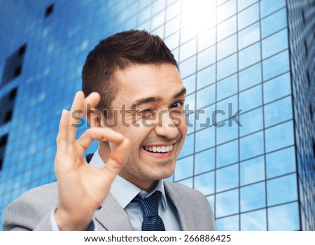 business, people, gesture and success concept - happy smiling businessman in suit showing ok hand sign over office building background