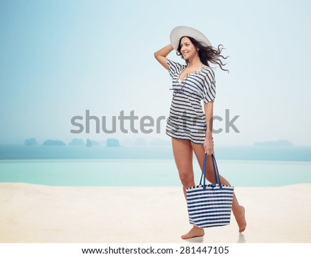 people, fashion, summer and beach concept - happy young woman in summer clothes and sun hat with bag over infinity pool at beach resort