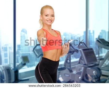sport, fitness, technology, gesture and people concept - smiling sporty woman smartphone showing thumbs up over gym machines background