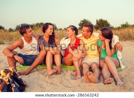 friendship, happiness, summer vacation, holidays and people concept - group of smiling friends sitting near fire on beach