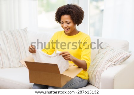 people, delivery, commerce, shipping and postal service concept - happy african american young woman taking clothes out of cardboard box or parcel at home