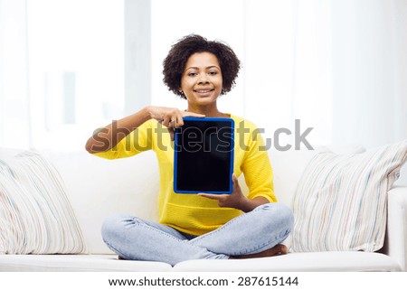 people, technology, advertisement and leisure concept - happy african american young woman sitting on sofa and showing tablet pc computer black blank screen at home