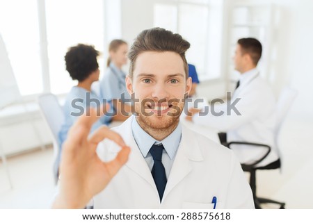 clinic, profession, gesture, people and medicine concept - happy male doctor showing ok hand sign over group of medics meeting at hospital
