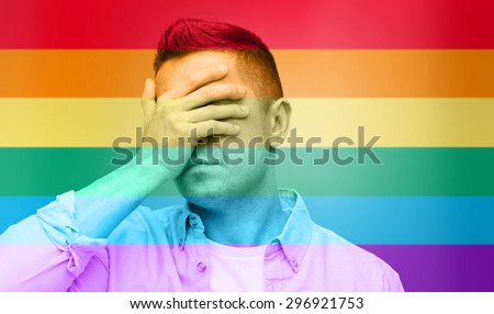 homosexual, homophobia, intolerance and people concept - unhappy gay man covering his eyes by hand over rainbow flag background