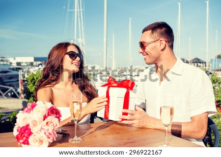 love, dating, people and holidays concept - smiling couple wearing sunglasses sitting with gift box, flowers and champagne at cafe