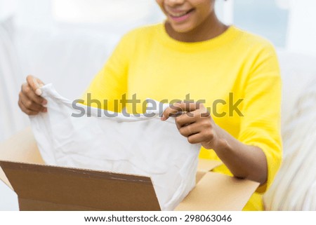 people, delivery, commerce, shipping and postal service concept - close up of happy african american young woman taking clothes out of cardboard box or parcel at home