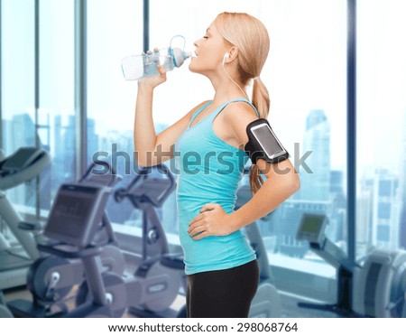 sport, fitness, technology and people concept - smiling sporty woman with smartphone and earphones listening to music and drinking water over gym machines background