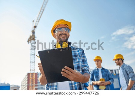 business, building, paperwork and people concept - happy builder in hardhat with clipboard and pencil over group of builders at construction site