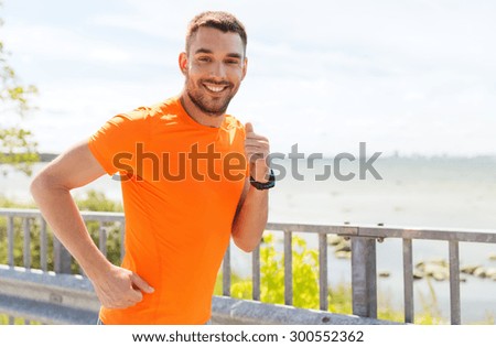 fitness, sport, people, technology and healthy lifestyle concept - smiling young man with heart rate watch running at summer seaside